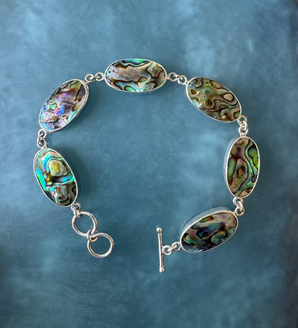 A handcrafted adjustable bracelet featuring multiple abalone shell bezels set in sterling silver. Each abalone piece glows with iridescent hues of blue, green, and violet, creating a luminous ocean-inspired design.